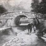 A black and white illustration of the Croydon Canal in the early 19th century. A small stone bridge goes over the canal, and a couple of people are walking alongside it.