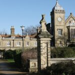 A photo of the Royal Waterman's Almshouses. Historic estate with stone statues at entrance, featuring a building with two towers, arched windows, and a mix of brick and stone architecture under a clear blue sky.